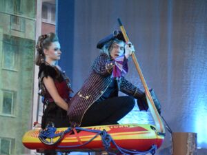 Ein rot-gelbes Schlauchboot. Darin zwei Schauspieler, Frau und Mann. Der Mann mit Ruder und Piraten Hut und Jacke, die Frau im Piratenkostüm.