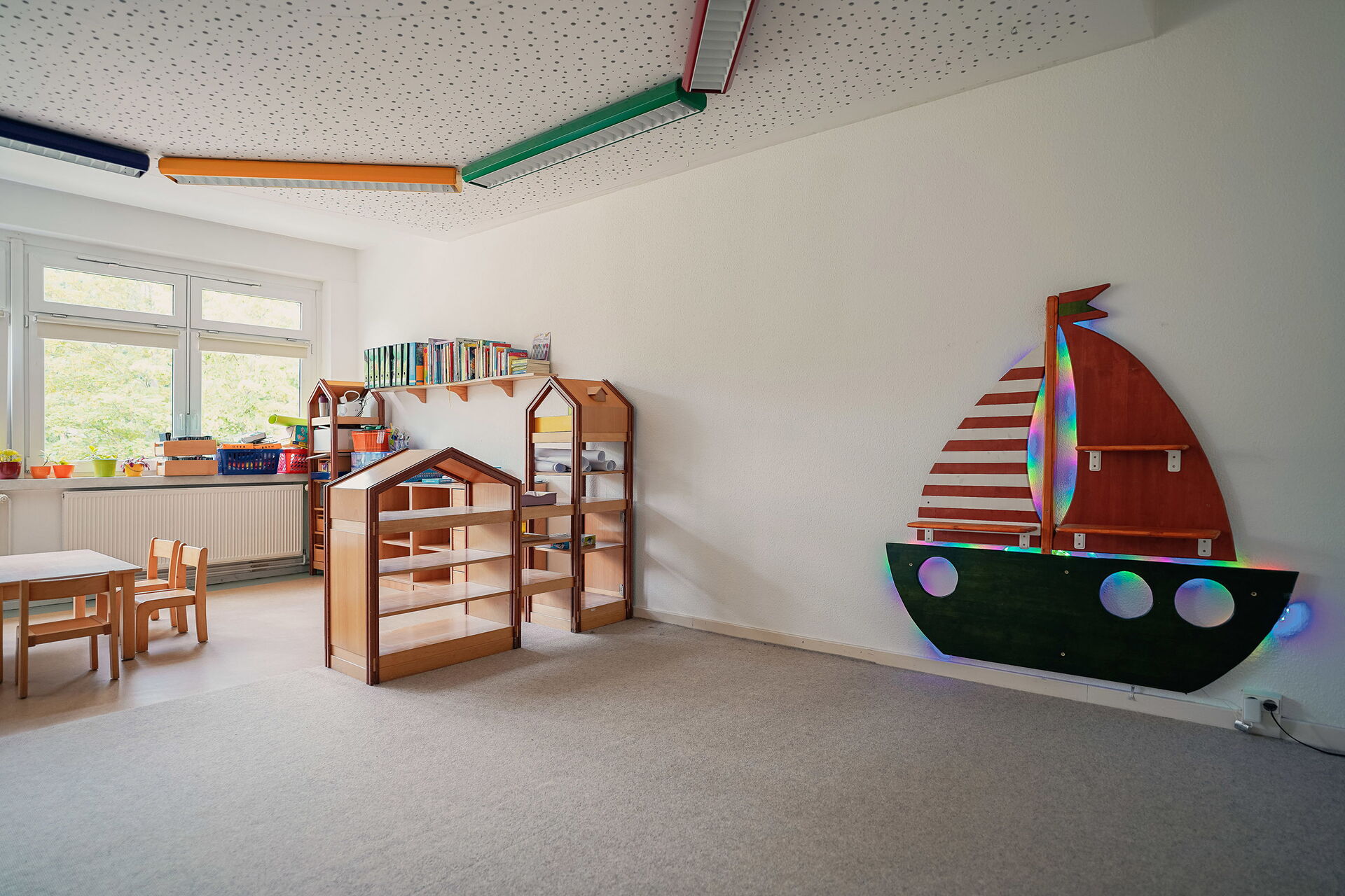 Großer heller Raum, hellgrauer Teppich im ganzen Raum. An der weißen Wand ein beleuchtetes Segelschiff aus Holz. Kleine Regale, eine Spielecke mit kleinem Tisch und Stühlchen.