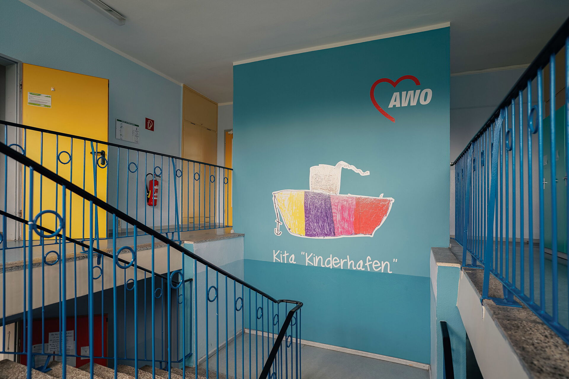 Treppenhaus in Blau. Auf einer Wand das AWO Logo und ein buntes Schiff mit Anker. Unter dem Schiff steht in weißer Schrift Kita "Kinderhafen".