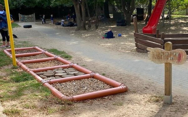 Spielplatz mit roter Rutsche im Vordergrund ein Barfußpfad. Kleine Kinder spielen auf dem Spielplatz.