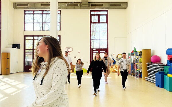 Eine Turnhalle  mit hellen Wänden. Junge Menschen schreiten durch die Halle. Kommen auf die Kamera zu.