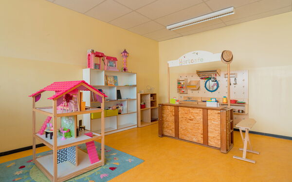 Spielzimmer. Puppenhaus mit pinken Dach auch einen geblümten kleinen Kinderteppich, Regal an der Wand mit Spielzeug. Und ein Spielzeug "Hort-Café" aus Holz mit Tresen und Ablagen für Geschirr dahinter an der Wand.