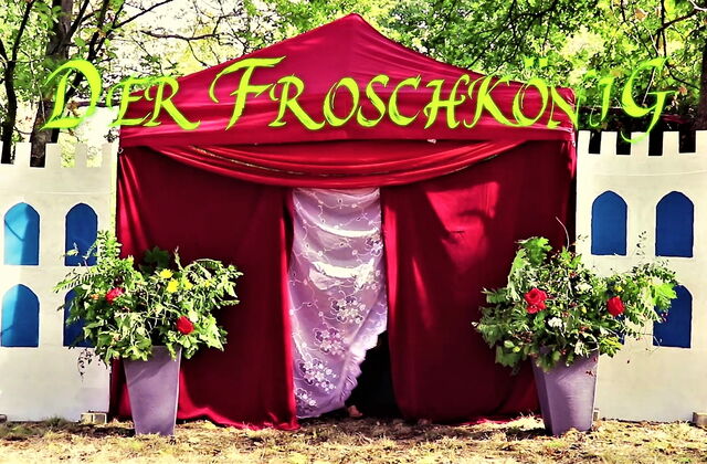 Draußen in der Natur. Ein rotes samtiges Märchenzeit, mit rosa Vorhang vor dem Eingang. Vor dem Zelt stehen links und rechts zwei Blumenkübeln. Schlossmauern aus Pappe sind links und rechts am Zelt.