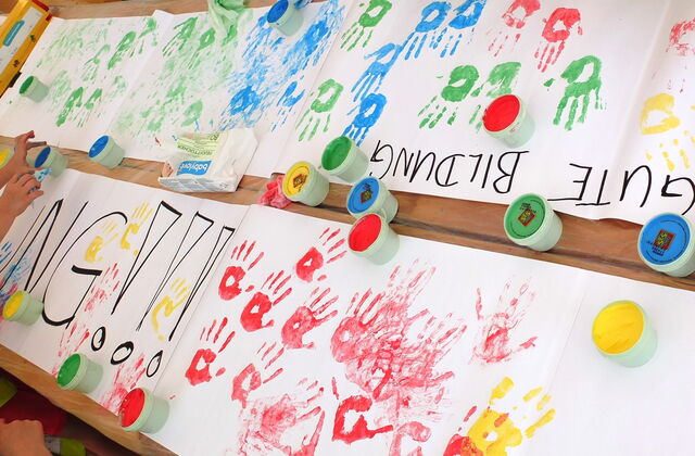 Großer Tisch, viele große Malblätter. Kinder haben ihre Hände in Farbe getunkt um Handabdrücke auf Papier zu bringen, es sind rote, gelbe, blaue, grüne Abdrücke. Text auf einem Blatt: GUTE BILDUNG