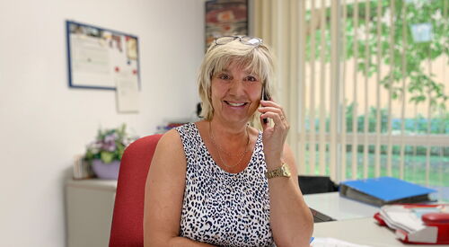 Frau an einem Streibtisch sitzend. Mit Telefon am Ohr. Frau ist blond, Brille auf den Kopf geschoben.
