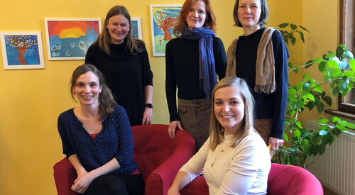 Fünf Frauen in einem Raum mit gelben Wänden und Kinderbildern an der Wand. Zwei Frauen sitzen in roten Sesseln, drei stehen dahinter. Alle lächeln freundlich in die Kamera.