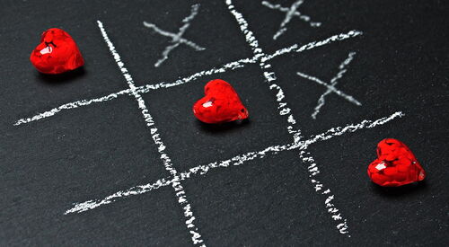 Tic-Tac-Toe-Spiel mit Kreide auf schwarzem Untergrund gezeichnet, drei rote Herzfiguren im Spielfeld.