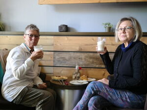 2 Frauen mit Getränken in der Hand. Sie sitzen an einem kleinen Tisch bei Kaffee und Kuchen, beide blicken in die Kamera.