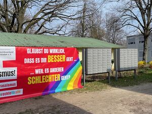 AWO Banner an einem Zaun, dahinter ein flaches Gebäude, davor ein Kiesweg. Auf dem AWO Banner steht: AWO gegen Rassismus! Glaubst Du wirklich, dass es Dir besser geht... ... weil es anderen schlechter geht? Auf dem Banner ist auch ein Regenbogen.