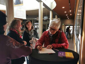 Kinofoyer. Stehtisch. Eine Frau mit grauen Haaren und Brille trägt sich in Liste ein.