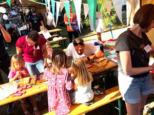 Es hängen bunte Wimpel und Luftballons rum. Kinder die an Biertischgarnituren sitzen und basteln und malen. 2 Erwachsene sitzen / stehen dabei.