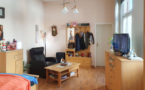 Bewohnerzimmer. Bick durch dem Raum zur Tür. Neben der Tür Garderobe, ein schwarzer Fernsehsessel, davor ein kleiner Couchtisch. Bewohntes Zimmer mit Kleidung und Deko.