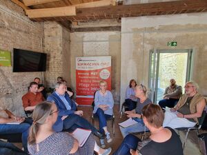 Menschen sitzen im Kreis auf Stühlen. In der Ecke des Raums steht ein Aufsteller der AWO. Eine Tür steht offen.