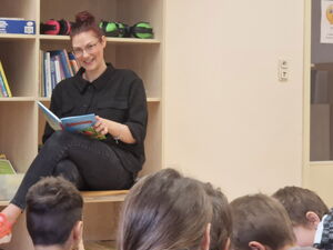 Eine Frau sitzt mit einem Buch in der Hand in einem Raum mit Regalen. Sie lächelt die Kinder, die vor ihr auf dem Boden sitzen an.
