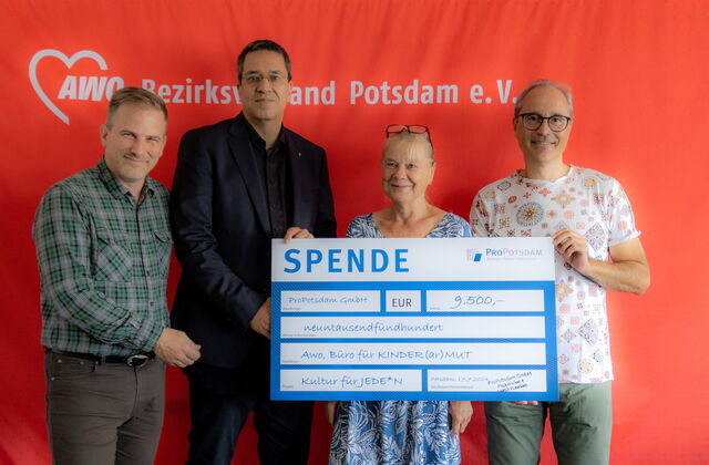 Vier Personen vor einer roten Wand mit AWO Logo und Schriftzug. Eine Frau und ein Mann halten einen großen Scheck in die Kamera. Zwei weitere Männer stehen links daneben. Alle vier lächeln in die Kamera.