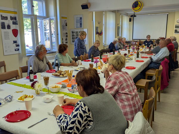 Die Lange Tafel schräg fotografiert. Menschen sitzen aber einige Stühle sind noch frei. Durch die offenen Fenster fällt Sonnenlicht.