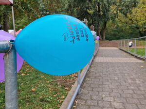 Ein blauer Luftballon mit der Aufschrift: „Kinder sind unsere Zukunft“ ist an einem Geländer befestigt. Dahinter sind weitere Ballons und ein gepflasterter Weg.