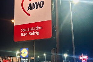 beleuchtetes Schild mit dem AWO Logo, im Hintergrund Straßenlaternen, Hauptstraße, beleuchtetes Schild von Lidl und Parkplatz von Rewe, Himmel ist dunkel