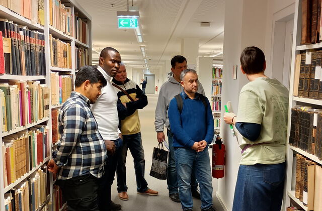 Teilnehmer werden durch die Bibliothek geführt. Sie stehen zwischen weißen Ragalen mit alten Büchern.