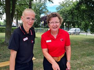 Draußen. Große rechteckige Torte wird von einer Frau mit Brille und rotem AWO Poloshirt angeschnitten. Auf dem Tisch stehen auch Teller und eine zweite schlankee Frau mit Brille und blonden Haaren und schwarzem AWO Poloshirt hält den Tortenheber bereit.