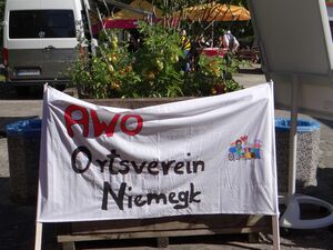 Draußen, Sonne scheint. Selbst bemaltes und beschriebenes Stoffbanner. Bild: kleine Gruppe von Menschen, Rollstuhlfahrerin, Kind mit Herz-Luftballon, alte Menschen, Mutter mit Baby auf dem Arm und Punkerin mit lila Haaren. Text: AWO Ortsverein Niemegk