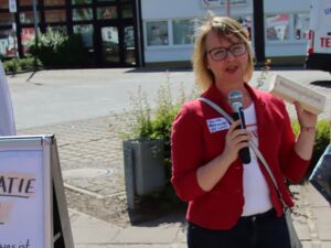 Im Freien. Frau mit Mikrofon in der Hand. Sie ist blond, trägt eine Brille, ein weißes T-Shit und darüber einen roten Blazer.