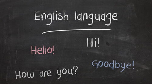 Eine dunkle Schultafel, an der mit bunter Kreide englische Wörter geschrieben sind. English language, Hello! Hi! How are you? Goodbye!