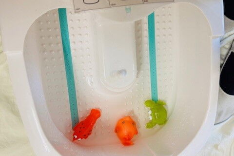 Ein Fußsprudelbad mit drei kleinen Quietschetieren. Davor hockt ein Kind mit weiteren Tieren neben sich. Auf dem Sprudelbad liegt eine Karte mit 6 Metacom Symbolen u.a. Wasser, Tiere, an, aus, rein und raus.