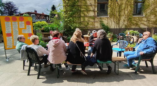 Gruppe von sitzenden Menschen um einen Tisch, darußen. Die Sonne scheint. Links eine Metaplan-Wand mit angehefteten Karten