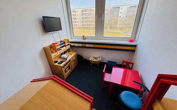 Ein Raum mit ausgestatteter Spielküche. Gegenüber steht ein roter Kindertisch mit zwei Stühlen, unter dem Fenster ein Puppenbett. An der Wand hängt ein Monitor.