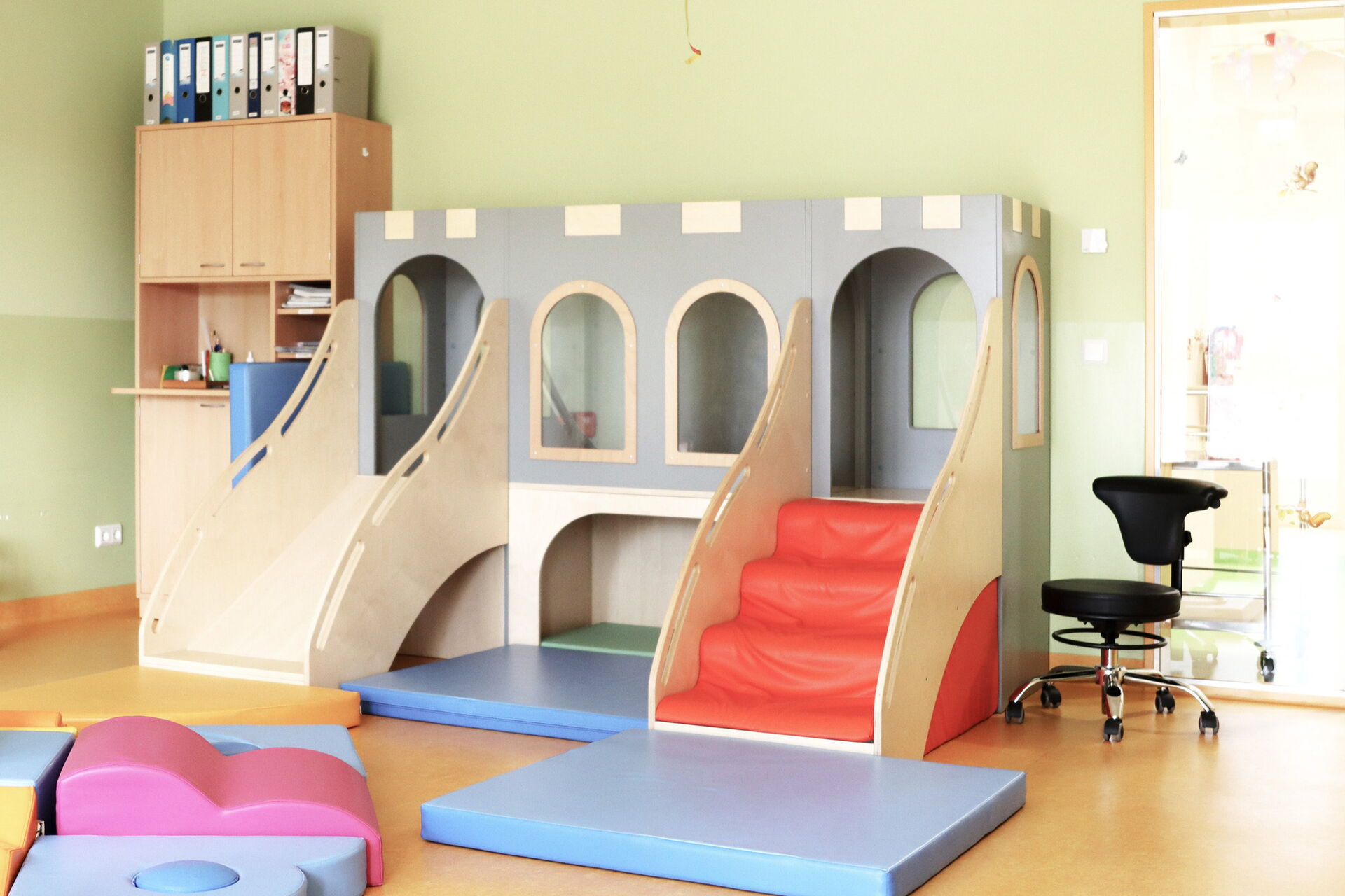Spielzimmer, Spiellandschaft. Mit roter Treppe auf der rechten Seite und Rutsche auf der linken Seite, sieht aus wie eine Burg. Daneben in der rechen Ecke steht ein hohes Regal mit Schranktüren.