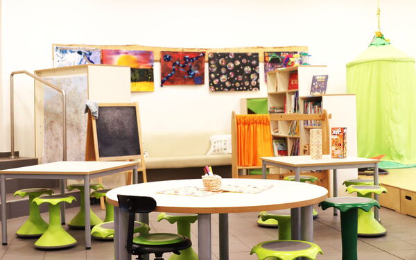 Ein Raum mit drei kleinen Tischen mit je 4 Hockern. Bilder hängen an der Wand. Kindertafel, grüne Hängehöhle und ein kleines Regal mit Büchern.