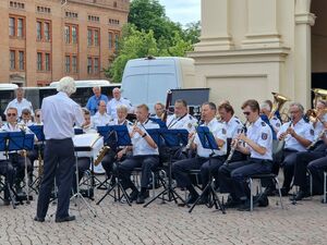 Auf einem Platz draußen. Das Polizeiorchester. Der Dirigent steht vor den sitzenden Ochestermitgliedern.