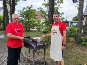 Zwei Männer am Grill. Beide tragen rote AWO T-Shirts. Einer hat eine weiße AWO Schürze darüber und kurze Hosen an. Der Grill ist leer und sauber.