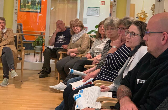 Großer Raum. 8 Menschen sitzen in einer Reihe. Stuhlreihe von der Seite fotografiert.