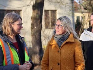 Draußen. 2 Frauen reden miteinander. Ein Mann steht rechts im Hintergrund.
