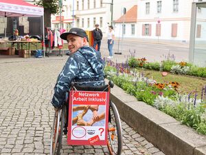 Draußen. Ein AWO Pavillon von weit. Rollstuhlfahrer im Aktiv-Rollstuhl von hinten, er dreht den Kopf und lächelt in die Kamera. Er trägt ein schwarzes Basecape und eine jacke. An seinem Rollstuhl ist hinten ein Plakat befestigt: Netzwerk Arbeit inklusiv.