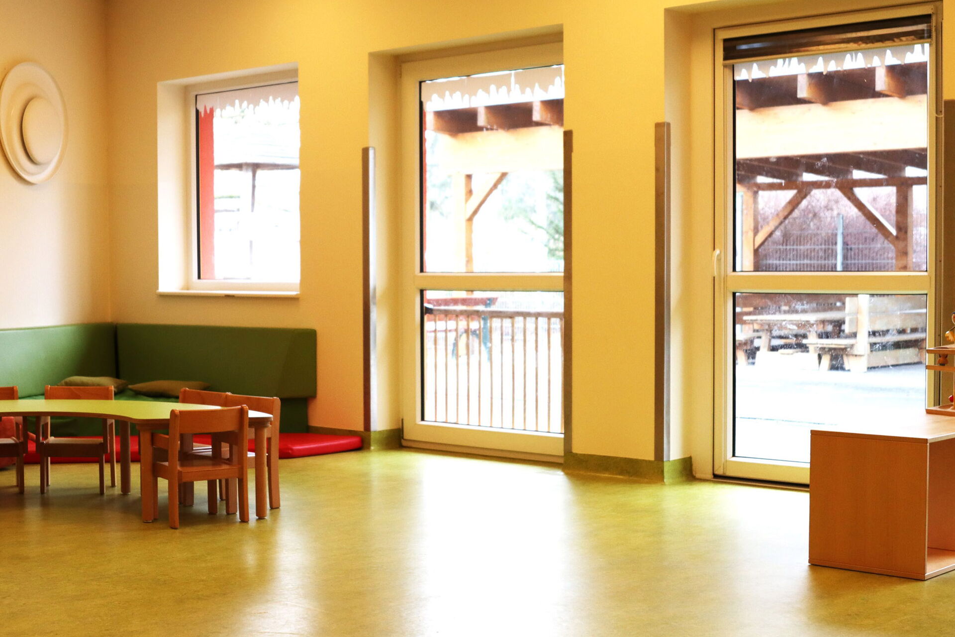 Heller Raum, kleine Tische und Stühle in der Ecke eine grüne Sitzecke. Rechts im Bild kleine Regale mit Spielzeug. Große Fenster lassen Licht hinein.