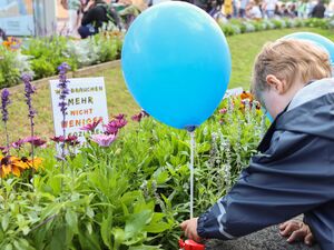 Blumenrabatte. Hinter die Blumen wurden zwei der Protestplakate gesteckt. Kleines Kind hat einen blauen SEKIZ Luftballon und steckt diesen in die Blumenrabatte.