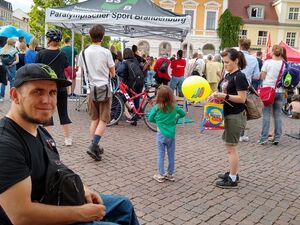 Blick über den Tisch mit Info-Material der AWO, ein Rollstuhlfahrer mit AWO T-Shirt und Base Cap lächelt in die Kamera. Fest Teilnehmende im Hintergrund. Ein Pavillon vom Paralympischen Sport Brandenburg.