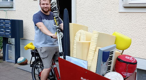 Mann mit T-Shirt und kurzer Hause sitzt auf Lastenfahrrad, der Laderaum ist voll beladen.