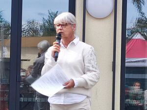 Frau vor einem Gebäude mit gelber Fassade und hohen Fenstern. Frau hat Mikrofon und eine DIN-A4 Seite in der Hand und häält eine Rede. Frau mit Brille, weißen Haaren und heller Kleidung.