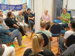 Großer Raum. Die Stühle sind in mehreren Kreisen angeordnet. Im inneren Kreis wird lebhaft diskutiert. An der Fensterwand hängen Schilder mit Forderungen der AWO.