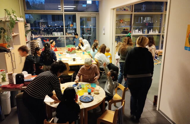 Raum, kleiner under Tisch und ein großer eckiger Tisch an beiden sitzen und stehen Erwachsene und Kinder. Ein Durchgang führt in einen anderen Raum.