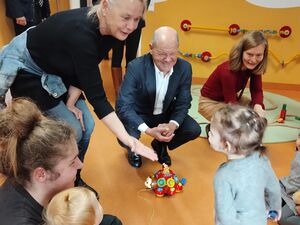 Ein kleines Mädchen tapst auf einen alten Mann mit Glatze im dunklen Anzug und weißem Hemd darunter zu. Eine Frau beugt sich vor streckt dem Mädchen die Hand entgegen. Eine Frau mit Kind auf dem Schoß sitzt auf dem Fußboden.