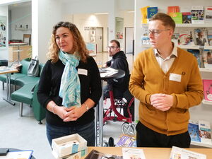 Frau und Mann stehen an einem Stand in einem Foyer. Auf dem Tisch vor ihnen liegt Info-Material.