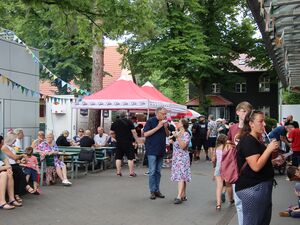 Blick aufs Gelände. Menschen sitzen an Picknicktischen, stehen am Essensstand an, gehen über das Gelände, unterhalten sich und sehen sich um.