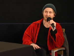 Mann sitzt auf einem Stuhl und spricht in ein Mikrofon. Der Mann trägt eine Mütze, eine rote Jacke über einem schwarzen Oberteil.