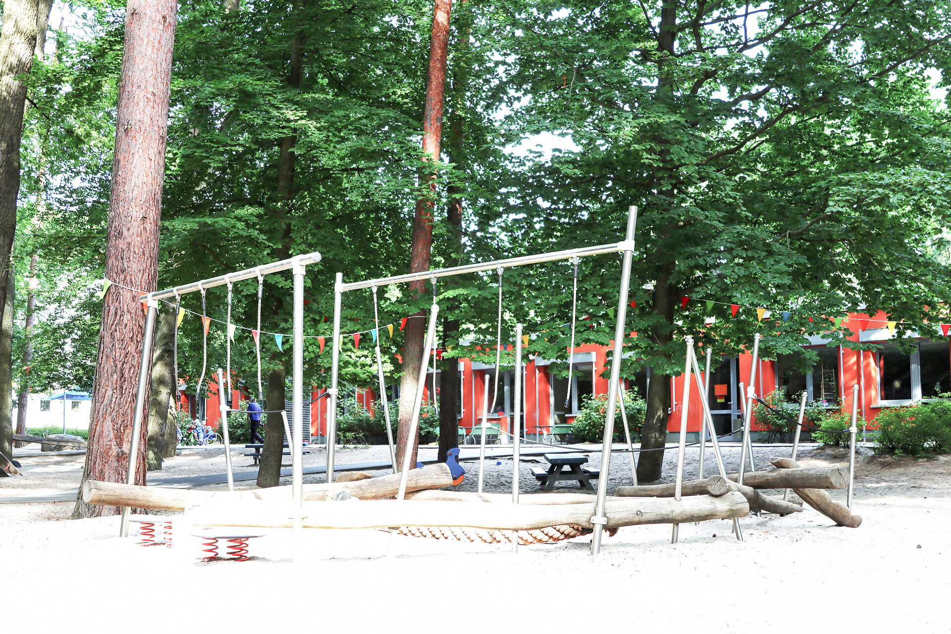 Spielplatz zwischen Bäumen. Durch die Bäume erkennt man das rote Kita Gebäude.