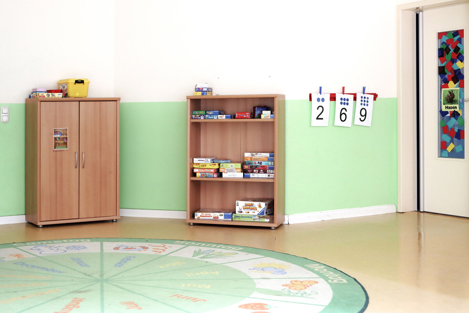 Spielzimmer. An der Wand ein offenes Regal mit Spielen. Auf dem Fußboben ein runder Kinderteppich mit den Wochentagen, und Monaten und dazu passenden Bildern.